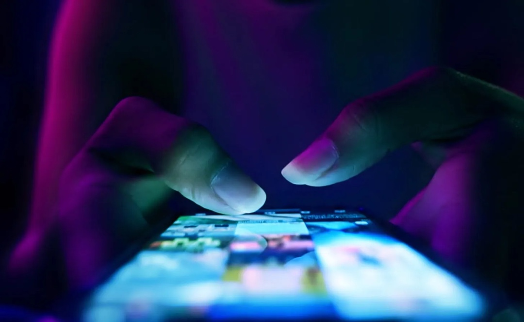 Hands scrolling through a brightly lit phone in a dark room.