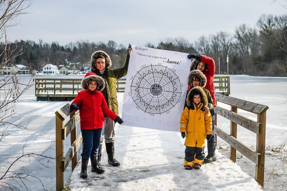 Children holding a large 1000 Hours Outside family tracker on a snowy bridge